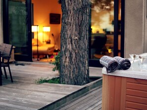 Une terrasse en bois avec un jacuzzi, un arbre et un intérieur chaleureux avec une lampe et un fauteuil.