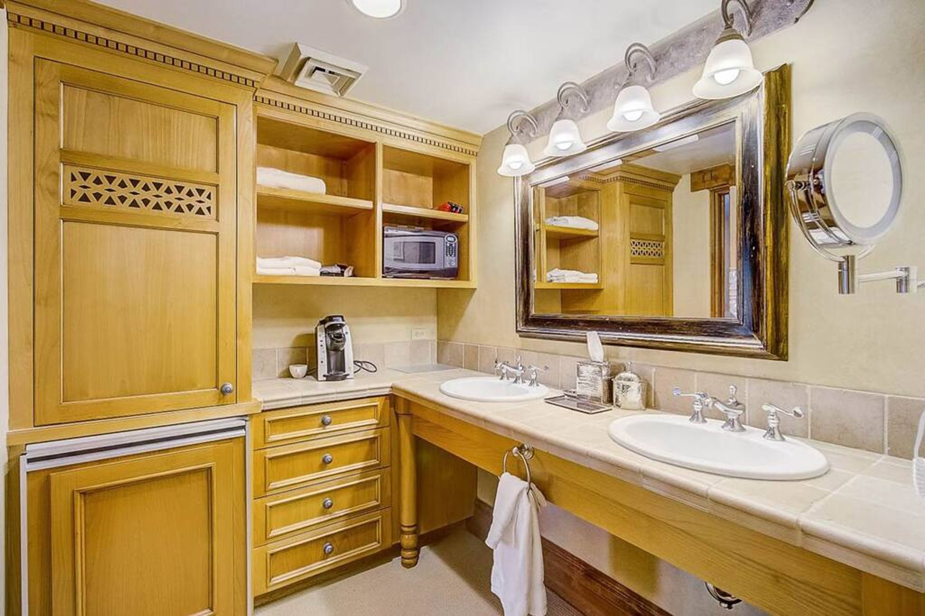 A bathroom with wooden cabinetry, two sinks, a large mirror with lights, and a microwave.