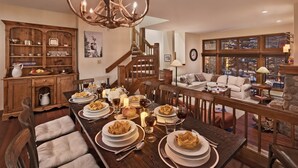 Une salle à manger avec une longue table en bois dressée pour le repas, un escalier menant à un niveau supérieur et un salon confortable doté d’une cheminée.