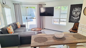A modern living room with a grey sofa, a coffee table, and a dining area with a wooden table and bar stools. - Dee-ones at Witboom With Pool (Cape Town)