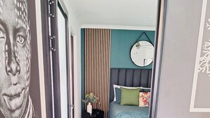 A bedroom with a bed, a bedside table, a chair, a mirror, and a wall with vertical blinds. - Dee-ones at Witboom With Pool (Cape Town)