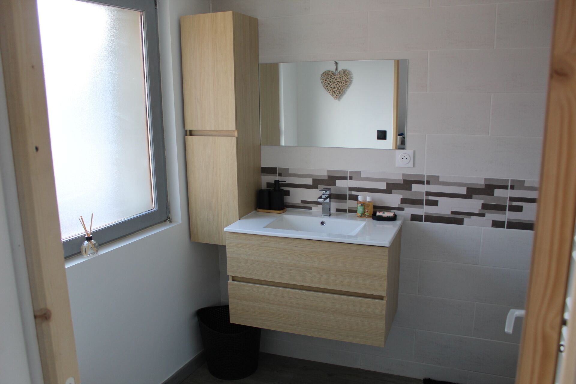 A bathroom with a white sink, wooden cabinets, a mirror with a heart decoration, and a tiled backsplash.