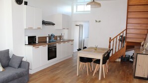 Une cuisine compacte avec des meubles blancs, un four à micro-ondes et une machine à café. On y trouve une table à manger avec des chaises, ainsi qu’un escalier menant à une mezzanine.