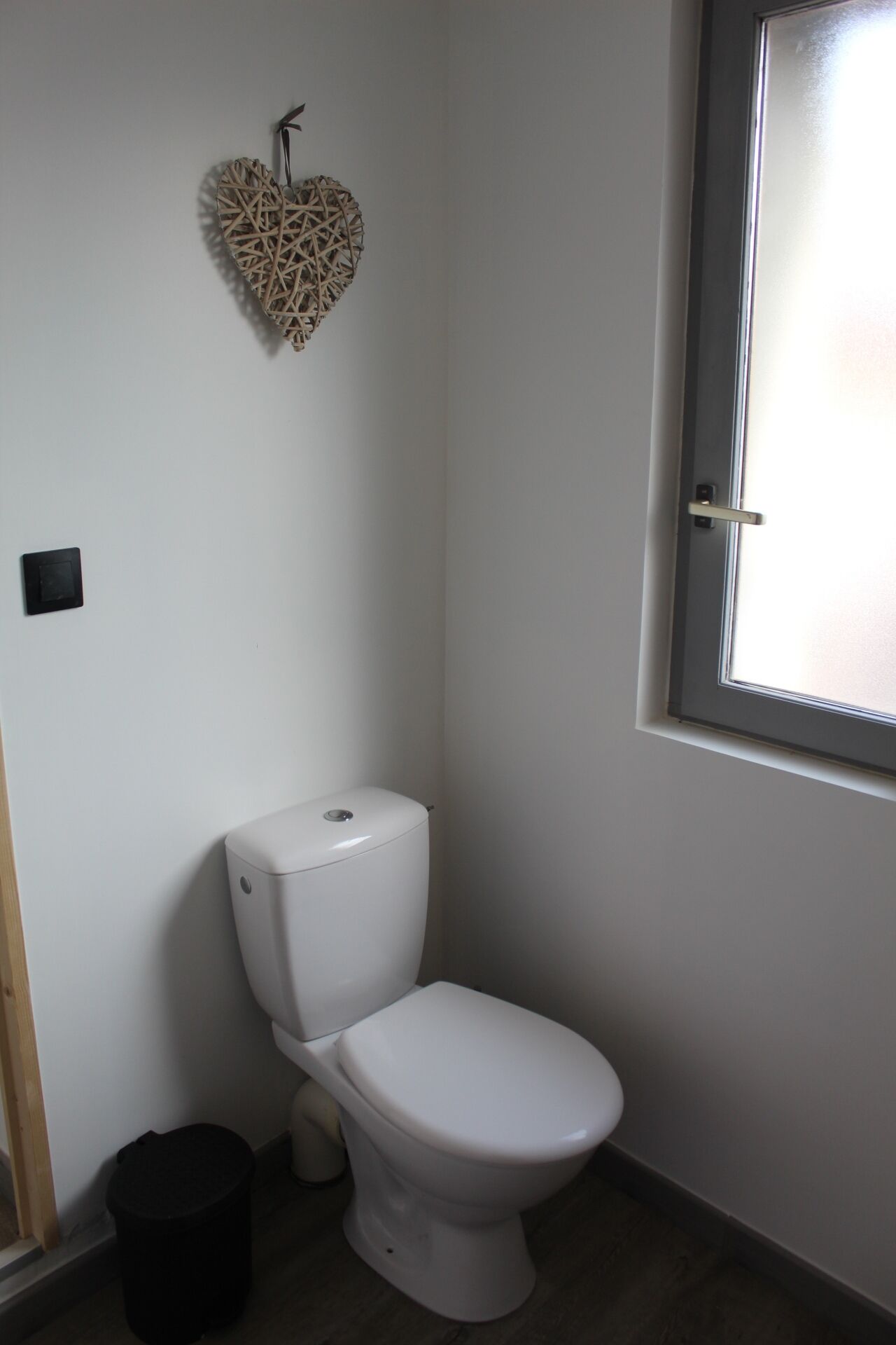 A small, white bathroom with a toilet, a black trash bin, and a window.