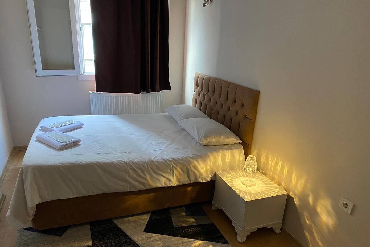 A neatly made bed with white linens and two pillows, a bedside table with a lamp, and a window with curtains.