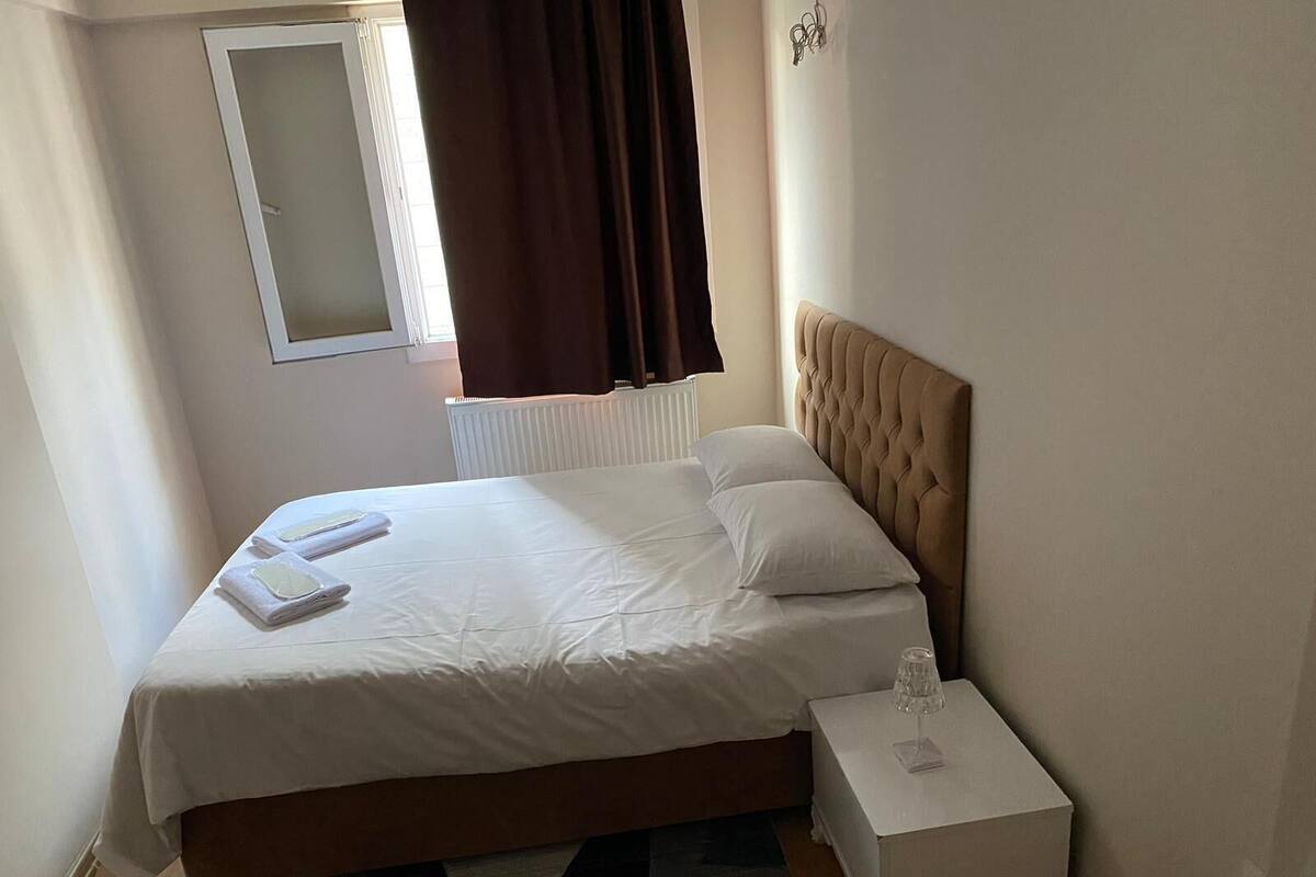 A neatly made bed with white linens, a bedside table with a glass, and a window with brown curtains.