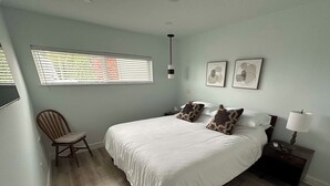 Un dormitorio con una cama grande, dos almohadas, una silla, una mesita de noche con lámpara y una ventana con persianas.