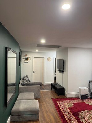 A modern living room with a grey sofa, a red patterned rug, a flat-screen TV mounted on the wall, and a white door leading to another room. - Cosy Entire House Open Kitchen 2 Bedroom for 5 (London)