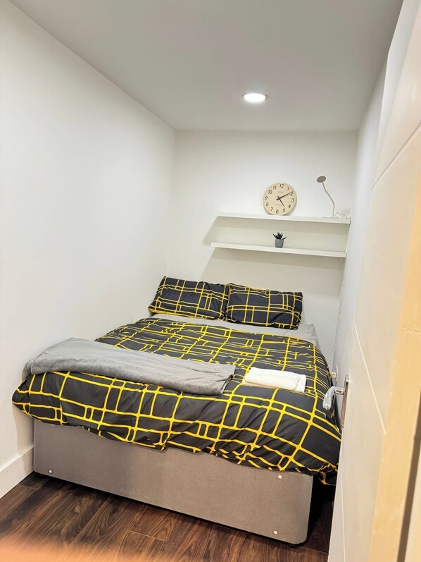 A small, neatly arranged bedroom with a bed, pillows, a clock, and a shelf with a plant. - Cosy Entire House Open Kitchen 2 Bedroom for 5 (London)