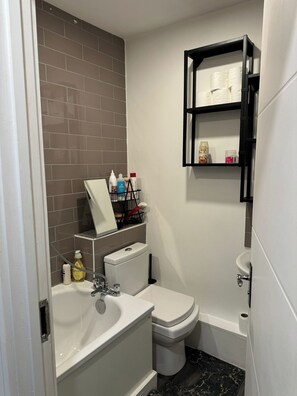A compact bathroom with a white sink, toilet, and bathtub. There is a shelf with toiletries and a towel rack above the toilet. - Cosy Entire House Open Kitchen 2 Bedroom for 5 (London)