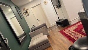 Een moderne woonkamer met een grijze bank, houten vloer, een rood tapijt met patroon, een grote spiegel en een wandklok.