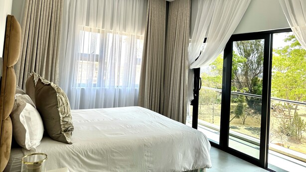 A modern bedroom with a large bed, sheer curtains, and a view of greenery outside. - The Cozy and Comfy Home In Portglen (Harare)