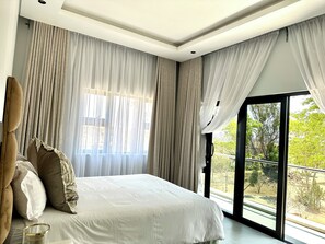 A modern bedroom with a large bed, sheer curtains, and a view of greenery outside.