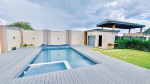 A rectangular swimming pool with a wooden deck, a small covered seating area, and a grassy lawn with a fence. - The Cozy and Comfy Home In Portglen (Harare)