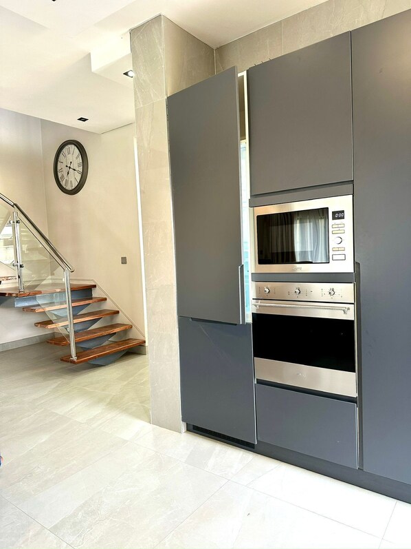 A modern kitchen with a built-in microwave and oven, a staircase with glass railings, and a clock on the wall.