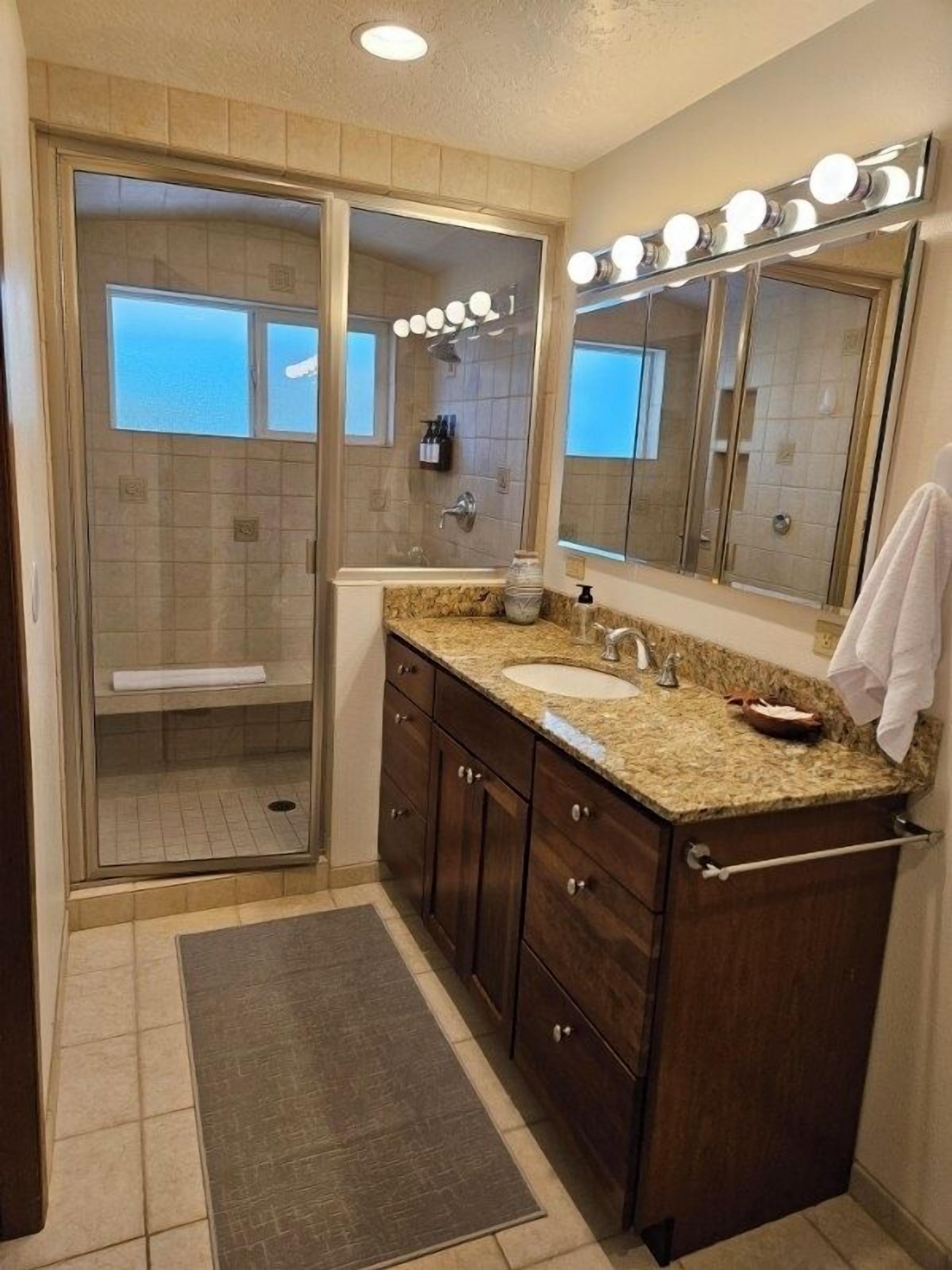 A bathroom with a glass-enclosed shower, a vanity with a sink, a mirror with lights, and a towel rack.