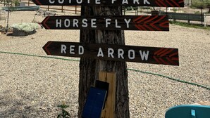 Un panneau indicateur directionnel avec de nombreuses flèches pointant vers différentes destinations, notamment Bath House, T-Down, Burnt Ridge, Coyote Park, Horse Fly et Red Arrow.