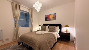 A neatly arranged bedroom with a bed, bedside tables, a window with curtains, a painting on the wall, and a chandelier.