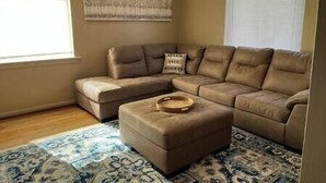 A living room with a sectional sofa, a small ottoman, a patterned rug, and a wall art piece. - Blue Suite at Brickhouse Inn (Hermann)