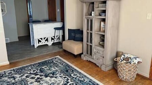A room with a wooden floor, a patterned rug, a white bar area, a cabinet with glass doors, and a wicker basket. - Blue Suite at Brickhouse Inn (Hermann)
