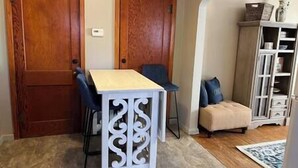 A room with a wooden door, a white table with a chair, a small ottoman, and a white cabinet with shelves. - Blue Suite at Brickhouse Inn (Hermann)