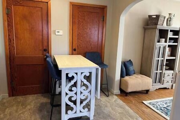 A room with a wooden door, a white table with a chair, a small ottoman, and a white cabinet with shelves. - Blue Suite at Brickhouse Inn (Hermann)