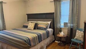 A bedroom with a bed, a bedside table, a chair, and a window with curtains. - Roam Suite at Brickhouse Inn (Hermann)