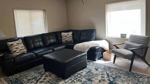 Una sala de estar con un sofá seccional de cuero negro, un otoman a juego, un sillón gris y un tapete estampado.