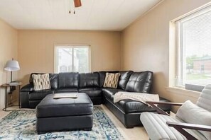 Un salon comprenant un canapé d’angle en cuir noir, un pouf assorti, un tapis à motifs, une table d’appoint avec une lampe et une fenêtre avec des stores.