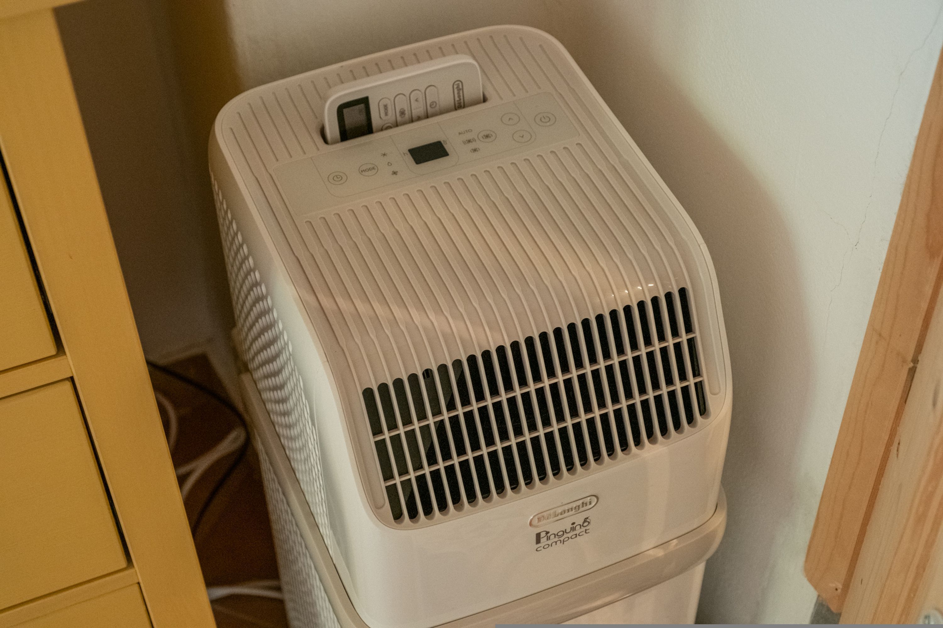 A compact, white portable air conditioner with a digital display and control panel, placed against a wall in a room with wooden cabinetry.