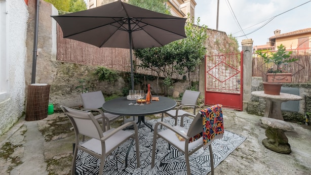 A round table with four chairs and a large umbrella in an outdoor patio area with a patterned rug, a stone table, and a decorative stone sculpture. - Aperol in Iseo With 1 Bedrooms and 1 Bathrooms (Iseo)