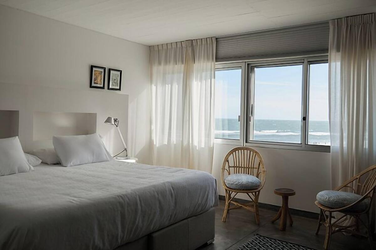 A bedroom with a large bed, two chairs, a bedside table, and a window with a view of the ocean.