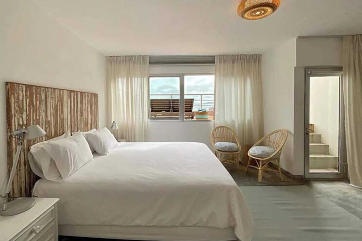 A bedroom with a large bed, two wicker chairs, a nightstand, a lamp, and a view of the sea through a window.
