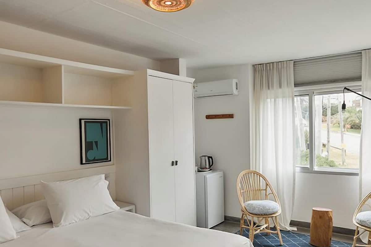 A hotel room with a bed, a wardrobe, a small kitchenette, two wicker chairs, a wooden stool, and a window with curtains.