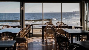 Un ristorante sulla spiaggia con tavoli e sedie in legno, ampie finestre che offrono vista sull'oceano e sullo skyline della città in lontananza.