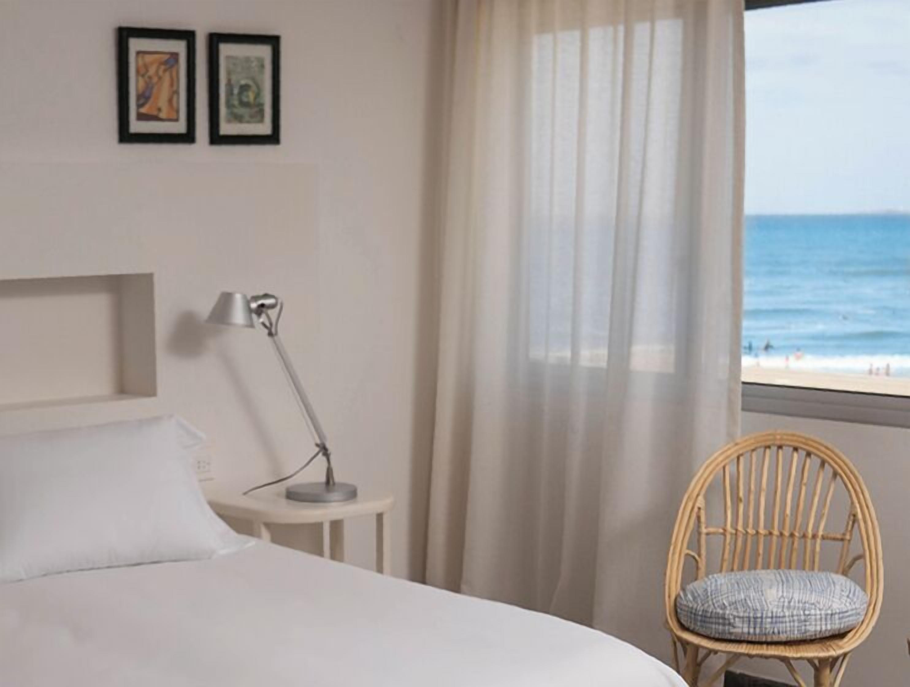 A hotel room with a bed, a bedside table with a lamp, a chair with a cushion, and a view of the beach through sheer curtains.