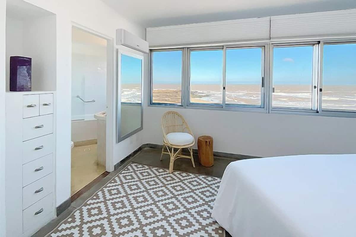 A hotel room with a bed, a chair, a wardrobe, a window with a beach view, and a patterned rug.