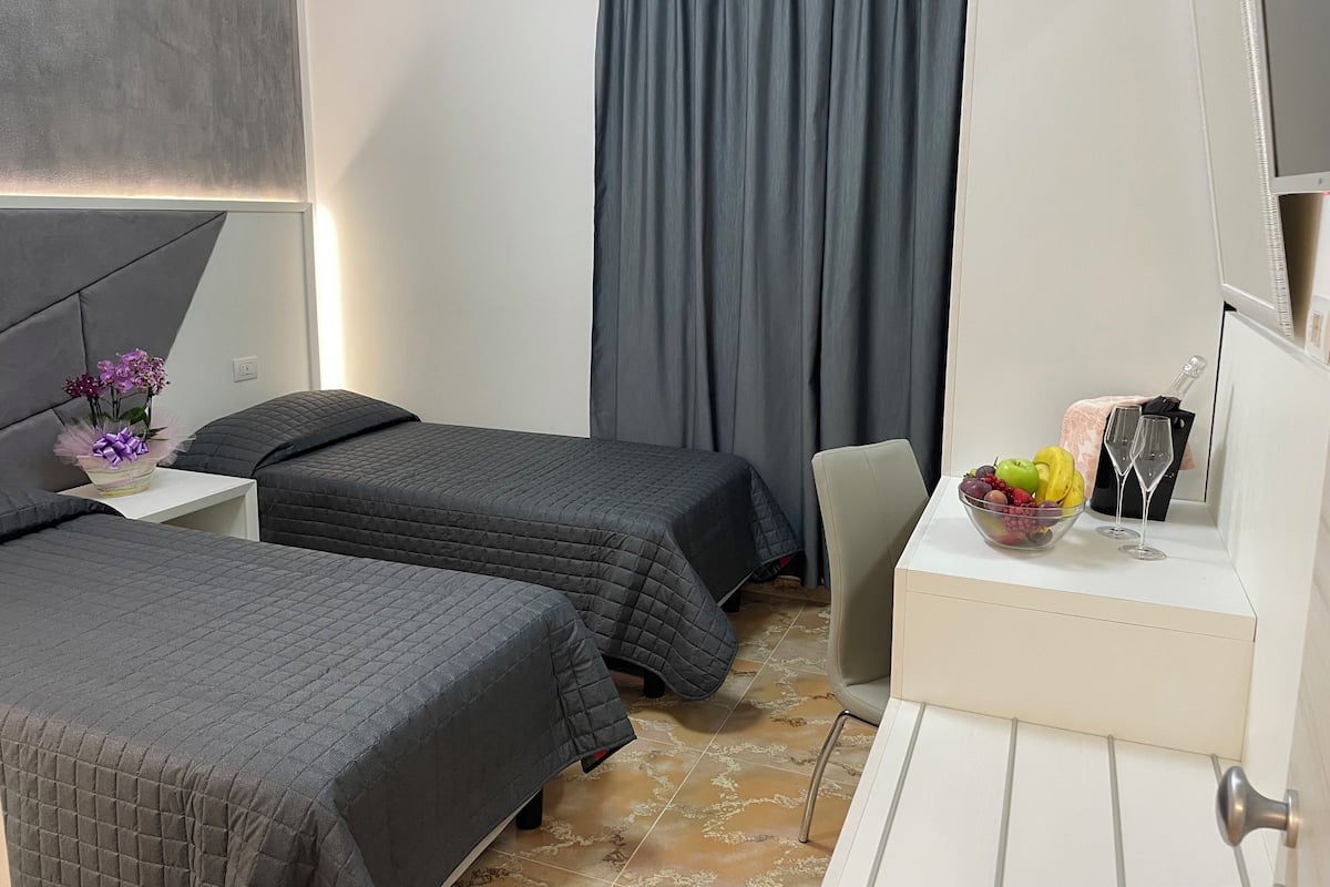 Hotelkamer met twee bedden, een bureau met stoel, een televisie, een gordijn en een fruitschaal met glazen.