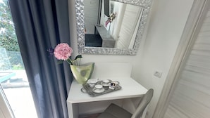 Un petit bureau blanc avec une chaise, un plateau contenant des tasses à thé et une théière, un vase avec des fleurs roses et un miroir orné d’un motif géométrique.