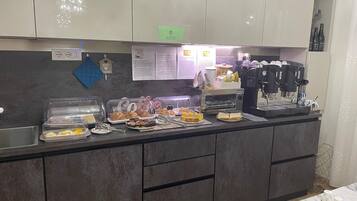 A breakfast bar with various food items, a coffee machine, and a sign indicating breakfast hours.