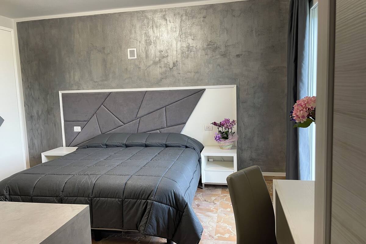 Een moderne slaapkamer met een groot bed, een witte nachtkastje, een klein tafeltje met een bloevas en uitzicht op de omgeving door een raam.