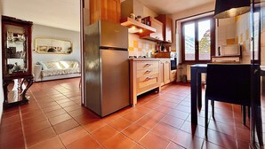 A kitchen with a refrigerator, wooden cabinets, a window, and a dining area with a chair. - Oh!megna in the historic heart (Omegna)