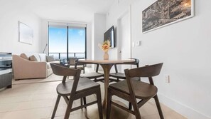 A modern living room with a round dining table, dark wooden chairs, a beige sofa, and a cityscape painting on the wall. - 49th Floor Sleek Condo | Cozy Pool, Gym & Spa Stay (Miami)