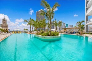 Une grande piscine extérieure bordée de palmiers, entourée de bâtiments modernes et surplombée d’un ciel bleu azur.