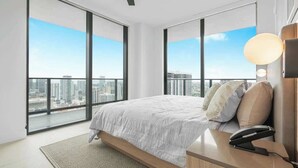 A modern bedroom with a large bed, a bedside table, a lamp, and a view of the city through large windows. - Lux 2BR Condo With Pool, Spa & Gym Access (Miami)