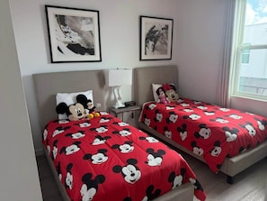 Two twin beds with red bedding featuring cartoon characters, a bedside table with a lamp, and framed artwork on the wall.
