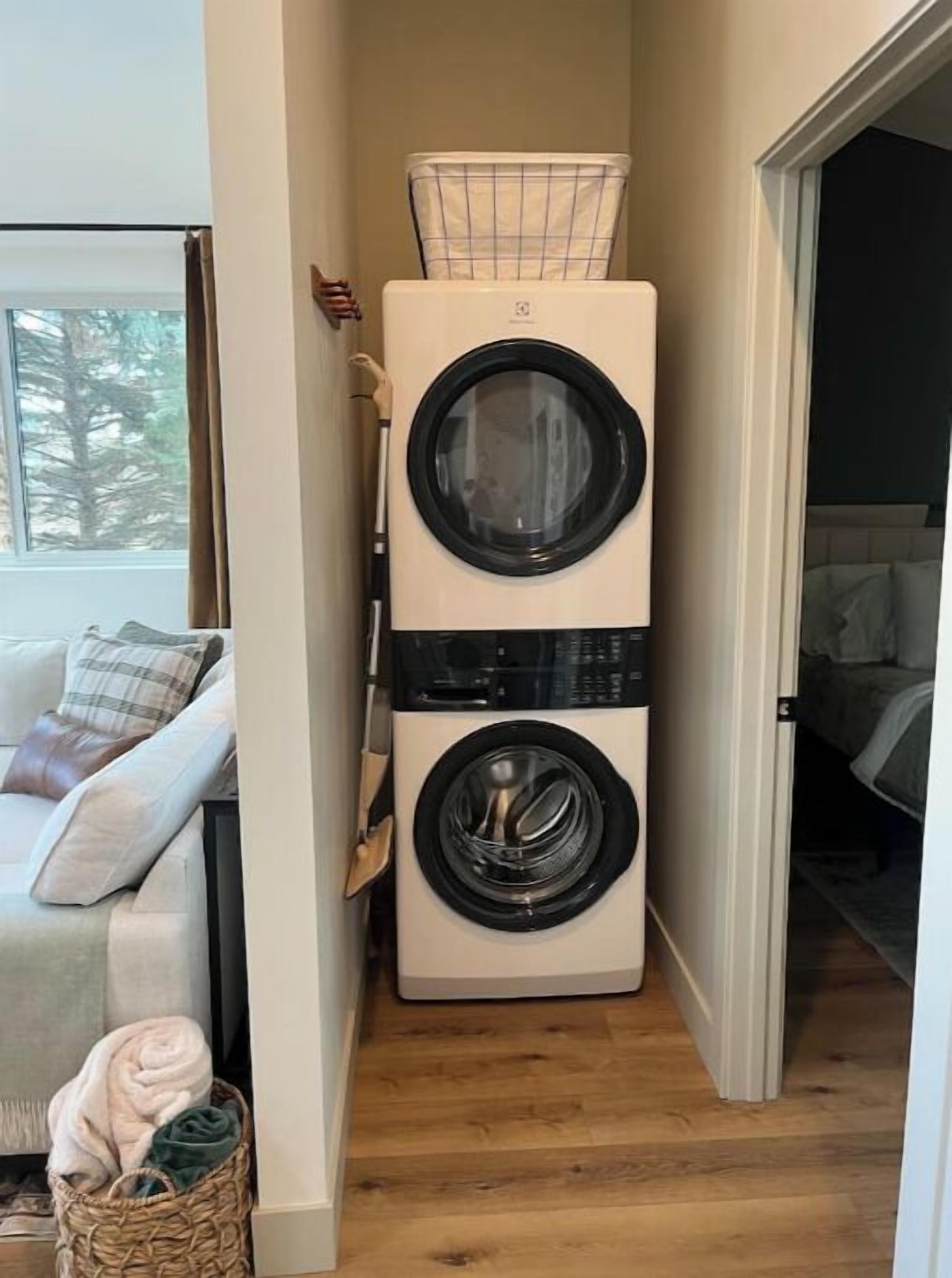 Een wasruimte met gestapelde wasmachines, een rieten mand met kleding en een uitzicht op een slaapkamer met een bed en een raam.