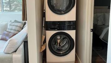 Een wasruimte met gestapelde wasmachines, een rieten mand met kleding en een uitzicht op een slaapkamer met een bed en een raam.