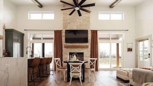 Un espacio habitable amplio con chimenea de piedra, una mesa de comedor con sillas, una zona de cocina con taburetes altos y un ventanal que proporciona luz natural.