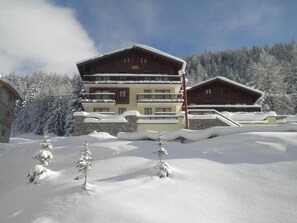 Un grand chalet à plusieurs étages, doté d’une façade en pierre et de niveaux supérieurs en bois, entouré d’arbres couverts de neige et surplombant un ciel dégagé.